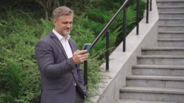 Takım elbiseli mutlu adam şehrin basamaklarında duruyor ve cep telefonunda daktilo yazıyor. İş, iletişim, teknoloji konsepti. Yavaş çekim