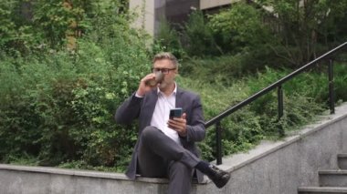 Kahve içerken ve dışarıda akıllı telefon kullanırken gülümseyen iş adamı dinleniyor. İş, iletişim, teknoloji konsepti. Yavaş çekim