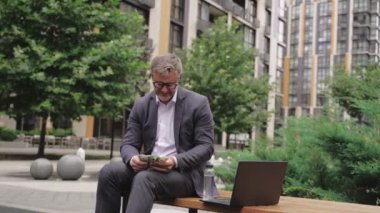Gözlüklü yetişkin işadamı bankta oturmuş para sayıyor ve dizüstü bilgisayarla çalışıyor. İş, ödeme konsepti. Yavaş çekim