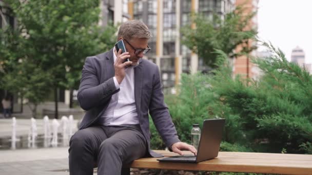 大人のビジネスマンは 携帯電話で話しながらラップトップにタイピングし ベンチに座っています ビジネス テクノロジーコンセプト スローモーション — ストック動画