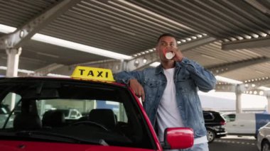 Genç taksi şoförü park yerinde kahve içerken kırmızı arabaya yaslanıyor. Sarı taksi arabası tabelası. İnsanlar, iş, hizmet kavramı. Yavaş çekim