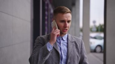 Tatminsiz genç iş adamı akıllı telefondan konuşuyor, dışarıda dikiliyor. İş, iş, teknoloji konsepti. Yavaş çekim