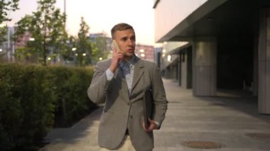 İşadamı cep telefonuyla konuşurken dizüstü bilgisayar tutuyor, cadde boyunca yürüyor. İş, iş, teknoloji konsepti. Yavaş çekim