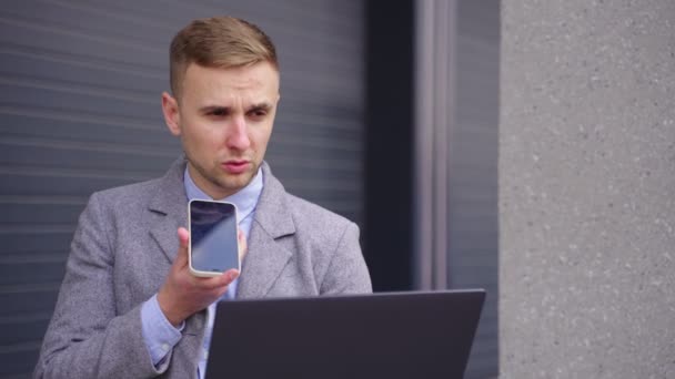 屋外でスマートフォンで話しながらラップトップで作業する若い男 ビジネス 技術コンセプト スローモーション — ストック動画
