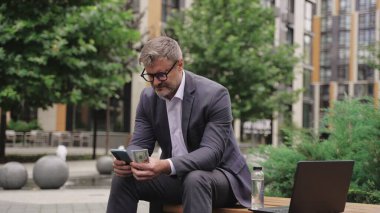 Gözlüklü bir işadamı elinde cep telefonu ve parayla bankta oturuyor. İş, ödeme, teknoloji konsepti. Yavaş çekim