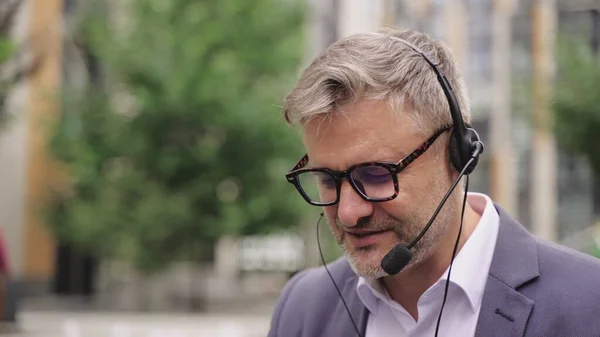 Adult businessman in glasses with headset sitting in the city outdoors. Business, technology concept. Slow motion