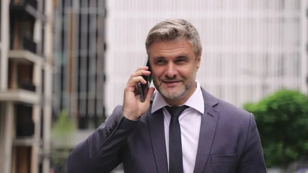 Smiling adult man in suite answering a phone call while walking along the street. Business, people, technology concept. Slow motion