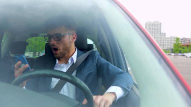 Takım elbiseli mutlu genç adam akıllı telefonla konuşurken şehirde araba kullanıyor. Ulaşım, iş, teknoloji ve insan duyguları kavramı. Yavaş çekim