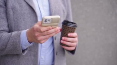 Açık havada duran bir iş adamı akıllı telefonuyla yazarken ve elinde bir fincan kahve tutarken. İş, teknoloji kavramı. Gerçek zamanlı.