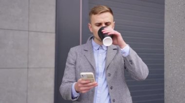 Genç adam kahve içiyor ve binanın yanında dikilirken akıllı telefon kullanıyor. İş, teknoloji kavramı. Gerçek zamanlı.
