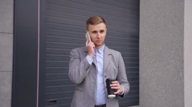 Gülümseyen genç adam cep telefonuyla konuşuyor, kahve içiyor, dışarıda dikiliyor. İş, teknoloji kavramı. Gerçek zamanlı.