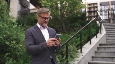 Gülümseyen yetişkin iş adamı elinde telefonla şehirde basamakların yakınında poz veriyor, yazı yazıyor. İş, iletişim, teknoloji konsepti. Yavaş çekim