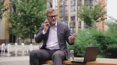 Yetişkin adam bankta oturuyor ve elinde para tutarken akıllı telefonla konuşuyor. İş, ödeme, teknoloji konsepti. Yavaş çekim