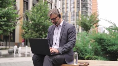 Kulaklık kullanırken dışarıda dizüstü bilgisayarla çalışan takım elbiseli, gülümseyen yetişkin bir adam. İş, teknoloji, insan kavramı. Yavaş çekim