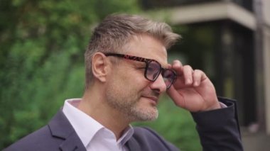 Gülümseyen işadamı sokakta durup gözlüğünü çıkarırken kameraya bakıyor. İş, insanların kavramı. Gerçek zamanlı konsept