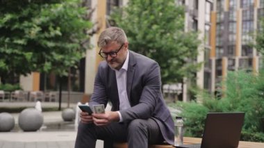 Gözlüklü bir işadamı elinde cep telefonu ve parayla bankta oturuyor. İş, ödeme, teknoloji konsepti. Gerçek zamanlı konsept