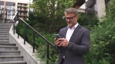Gülümseyen yetişkin iş adamı elinde telefonla şehirde basamakların yakınında poz veriyor, yazı yazıyor. İş, iletişim, teknoloji konsepti. Gerçek zamanlı konsept