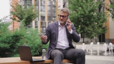 Yetişkin adam bankta oturuyor ve elinde para tutarken akıllı telefonla konuşuyor. İş, ödeme, teknoloji konsepti. Gerçek zamanlı konsept