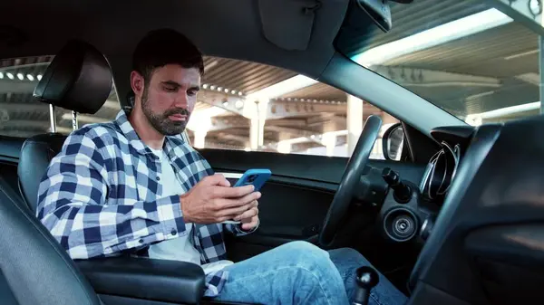 Guy sitting in the car while holding smartphone, typing, drinking coffee. Transport, technology, lifestyle concept