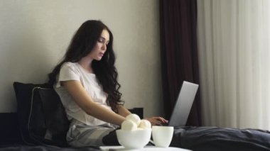 Dizüstü bilgisayar kullanan, yatakta otururken yazan bir kadın. Ağır çekim. Ev konseptinde çalışmak. Merkez Ofis