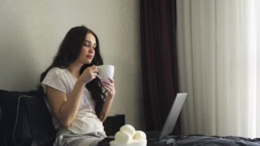 Evden bilgisayarla çalışan, kahve içen, yatakta oturan serbest çalışan bir kadın. Ağır çekim. Ev konseptinde çalışmak. Merkez Ofis