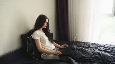 Güzel, genç bir bayan internette çalışırken, yatak odasında otururken eğleniyor. Ağır çekim. Ev konseptinde çalışmak. Merkez Ofis