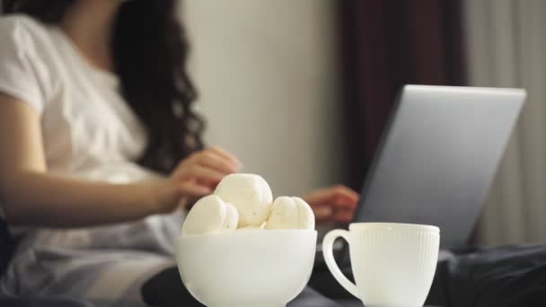 年轻的女士坐在床上 一边吃棉花糖一边在笔记本电脑上打字 慢动作在家工作的概念 总部办公室 — 图库视频影像