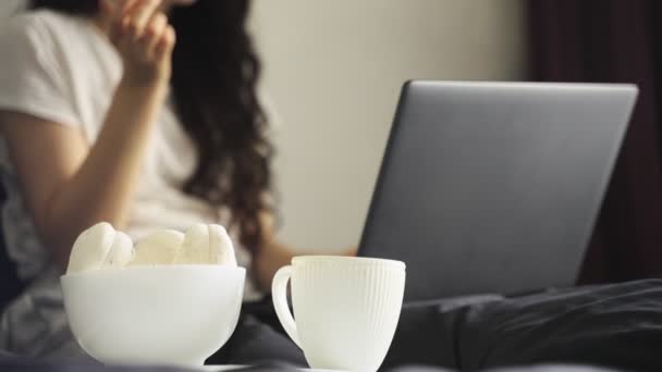 女人坐在床上 一边在笔记本电脑上打字 一边吃棉花糖 慢动作在家工作的概念 总部办公室 — 图库视频影像