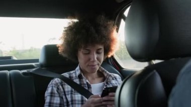 Akıllı telefon kullanan ve arabanın arka koltuğunda mesaj yazan gülümseyen genç bir kadın. İnsanlar, yaşam tarzı, teknoloji konsepti. Yavaş çekim