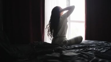 Yatak odasında yatakta oturup güneşin tadını çıkarırken pencereden dışarı bakan güzel bir kadın. Ağır çekim. Yaşam tarzı kavramı