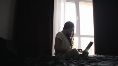 Pencerenin yanındaki yatak odasında oturan ve evdeki laptopta sohbet eden bir kadın. Ağır çekim. Yaşam tarzı kavramı