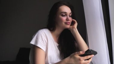 Akıllı telefon kullanarak ve pencerenin yanında oturarak evde dinlenen güzel genç bir kadın. Ağır çekim. Yaşam tarzı kavramı