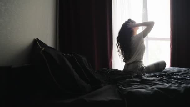 ベッドの上に座っている若い女性は 自宅でカーリーヘアに触れながら窓の外を見ていました スローモーション ライフスタイルコンセプト — ストック動画