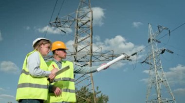 Elektrik hattı müfettişi panoya yazı yazarken meslektaşı elinde planlarla uzaktaki bir şeyi işaret ediyor. Yavaş çekim