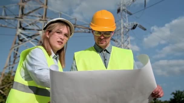 経験豊富なエネルギー監査人と彼女の同僚は 現場検査中に電気パイロンの近くに立っている間 青写真を読んでいます スローモーション — ストック動画