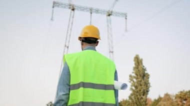 Kolunun altında çizimler olan koruyucu miğferli güç hattı müfettişi şanzıman kulesine doğru yürüyor. Yavaş çekim