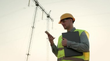 Elektrik direğinin yanında akıllı telefondan konuşurken taşınabilir bilgisayar tutan memnun enerji denetçisinin belden yukarı portresi. Yavaş çekim