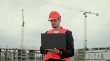 Yelekli, koruyucu kasklı ve gözlüklü bir erkek olay yerinde duruyor ve dizüstü bilgisayarda yazıyor. Yavaş çekim