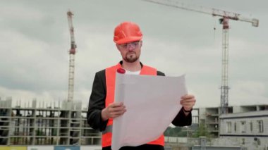 Koruyucu miğfer, yelek ve gözlüklü genç ustabaşı elinde büyük planlar ve çalışmalar tutuyor. Yavaş çekim