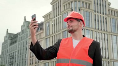 Korumalı yelek giyen profesyonel müteahhit sahada çalışıyor, akıllı telefon tutuyor ve selfie çekiyor. Yavaş çekim