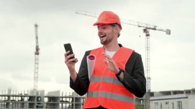 Koruyucu yelek giyen profesyonel bir adam şantiyede çalışıyor, elinde akıllı telefon tutuyor ve internetten selam veriyor. Yavaş çekim