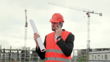 Koruyucu yelek giyen inşaatçı sahada çalışıyor, akıllı telefon tutuyor ve üzgün, kızgın duygularla konuşuyor. Yavaş çekim