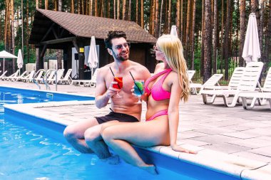Mutlu çift açık havuzun kenarında otururken kokteyllerin tadını çıkarıyor. Yaz tatili, aktif eğlence, yaşam tarzı konsepti.