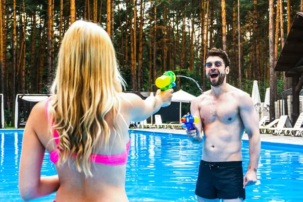 Yüzme havuzunda kalıp su tabancalarıyla oynayıp eğlenen sevimli bir çift. Yaz tatili, aktif eğlence, yaşam tarzı konsepti.