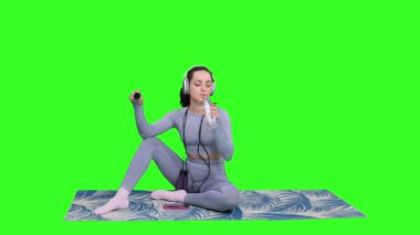 Kulaklıklı, zıplama halatıyla yoga minderinde oturan yeşil izole edilmiş arka planda dinlenen genç bir kadın. Fitness, spor, sağlıklı konsept