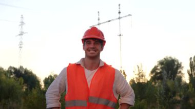 Kasklı gülen elektrik mühendisi yüksek voltajlı elektrik hatlarının önünde dışarıda poz veriyor. Dağıtım ve elektrik tedariği. Enerji endüstrisi konsepti. Yavaş çekim