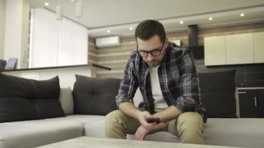 Mutsuz sakallı genç adam evde yalnız başına otururken akıllı telefon kullanıyor. Yalnızlık, depresyon, duygu konsepti. Yavaş çekim