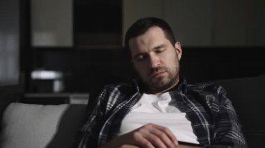 Evdeki modern oturma odasında yorgun ve sakallı bir adam rahat bir kanepede uyuyor. Dinlenme, aşırı çalışma, yorgunluk konsepti. Yavaş çekim