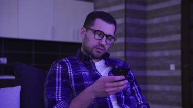 Gözlüklü, ciddi bir genç adam evdeki koltukta oturmuş telefonla sohbet ediyor. Yaşam tarzı, duygular, teknoloji konsepti. Yavaş çekim