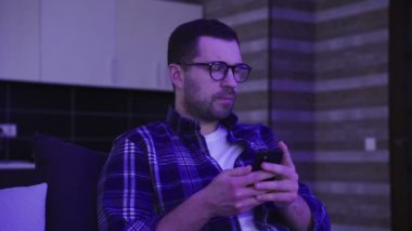 Mesajı okurken, gözlüğünü çıkarırken, evdeki kanepede otururken akıllı telefon kullanan endişeli genç adam. Yaşam tarzı, duygular, teknoloji konsepti. Yavaş çekim